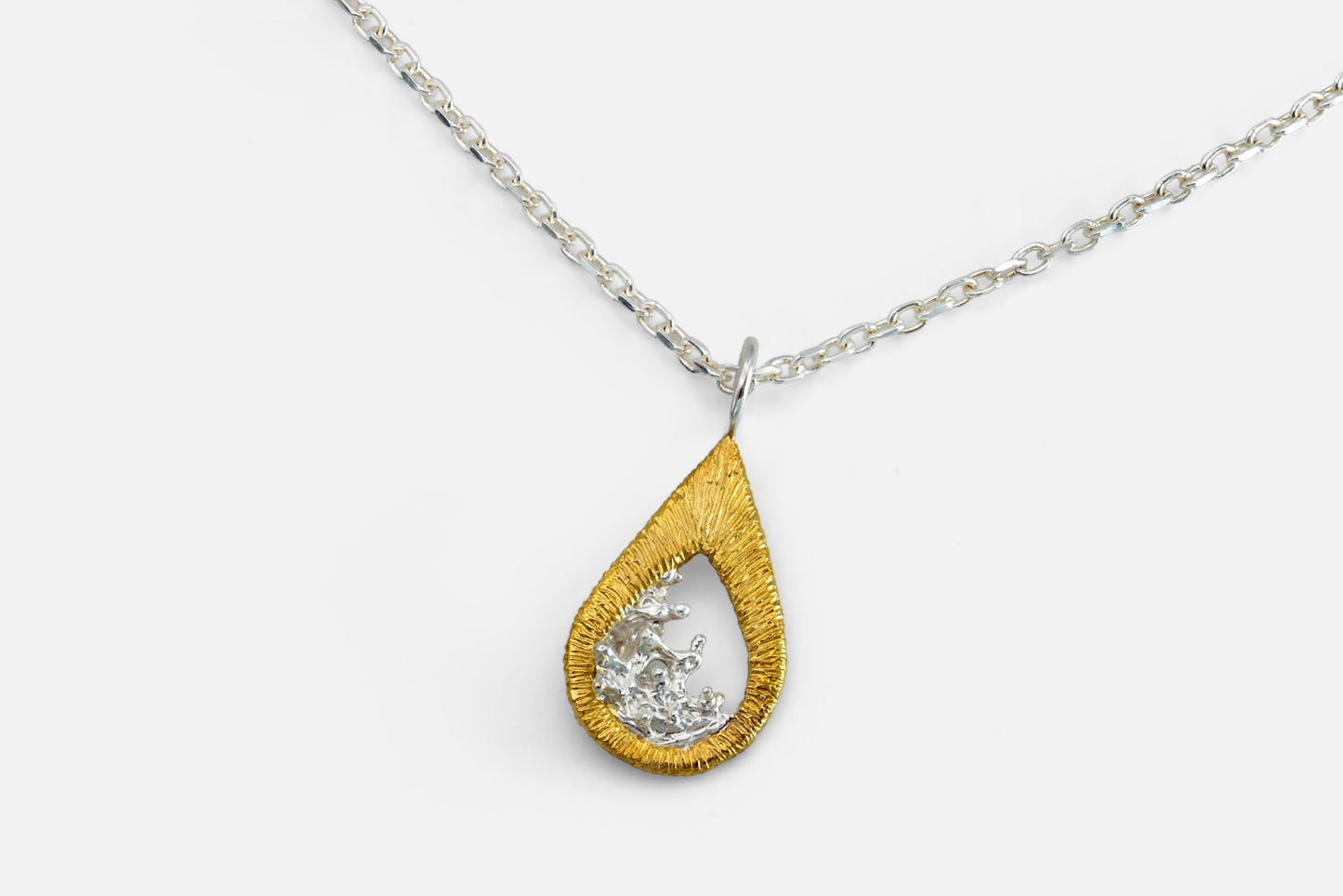 A teardrop-shaped gold vermeil and silver pendant necklace featuring a textured gold outer frame and a silver-toned inner design shaped like moving water, suspended from a silver chain against a light gray background.