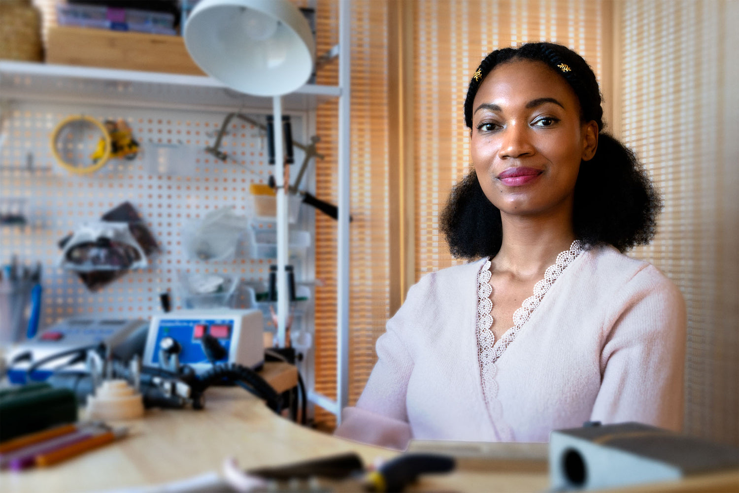 A woman with dark curly hair in a ponytail sits at a desk in a workshop or studio, wearing a pale pink top with a lace V-neckline. The background features a white pegboard with tools, a desk lamp, and beige curtains.
