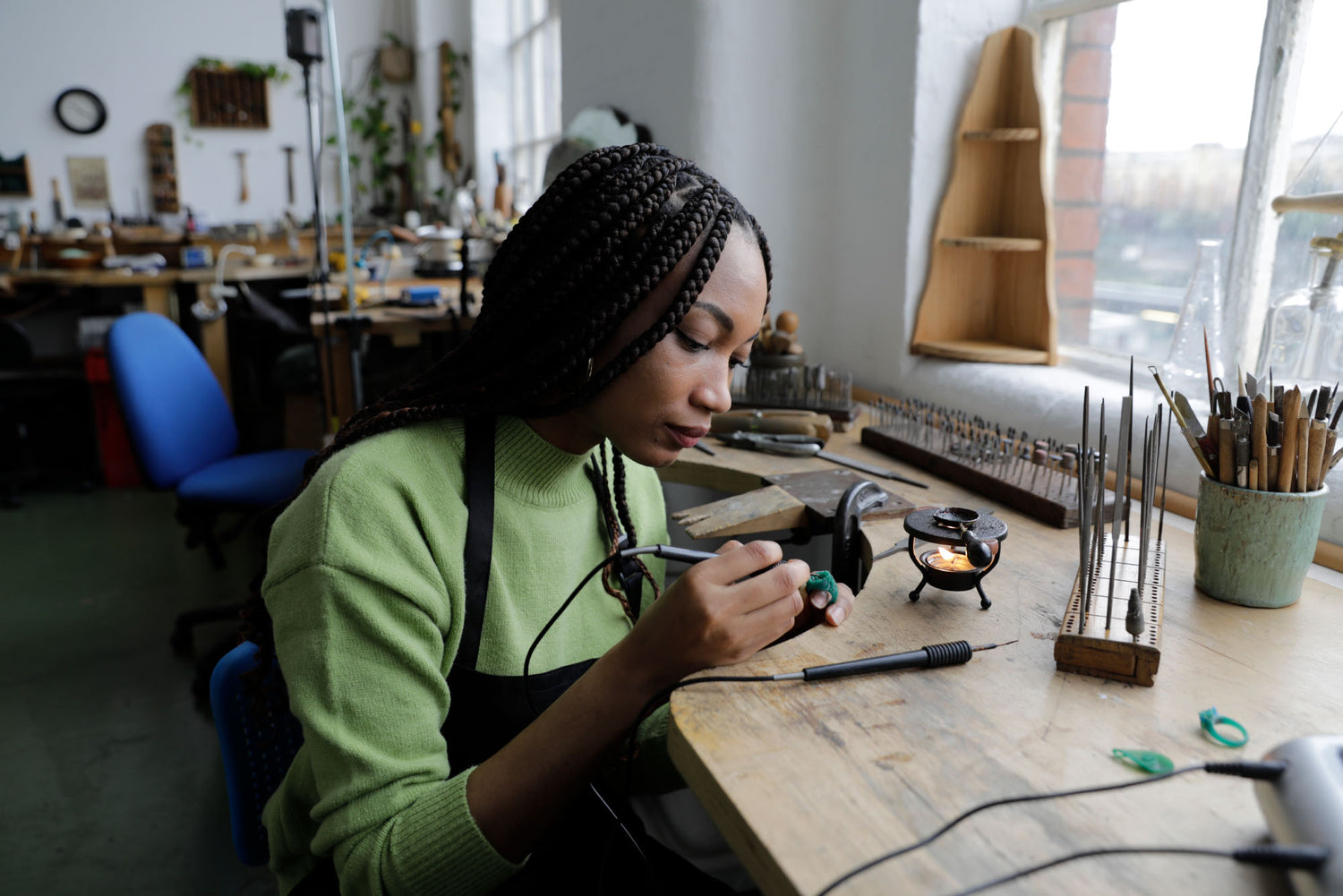 A woman wearing a light green sweater and black apron works at a wooden craft table in a bright workshop, using a small soldering tool. The table is filled with jewelry-making supplies, including a small soldering station, metal rods, and a cup of brushes.