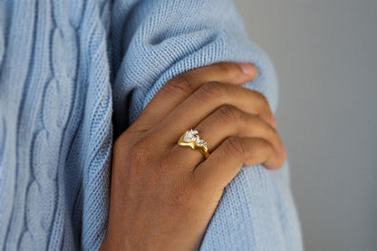 A close-up of a person's hand wearing a gold vermeil and silver ring with a wavy, organic shape and a splash of water crafted in silver in the center, resting on a light blue cable-knit sweater against a soft gray background.