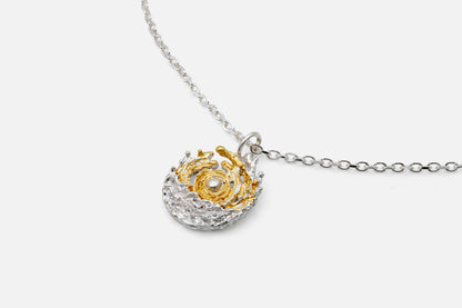 A delicate silver chain necklace featuring a spherical pendant with a gold swirl around a central sun, set against a textured silver background like the sea and tides moving at dawn, shown against a light gray background.
