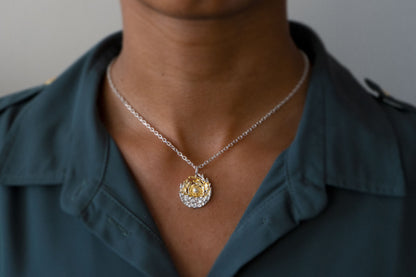 A close-up of a person wearing a delicate silver chain necklace with a round pendant featuring a gold swirl around a central sun, set against a textured silver background like the sea and tides moving at dawn, styled with a dark teal collared shirt.
