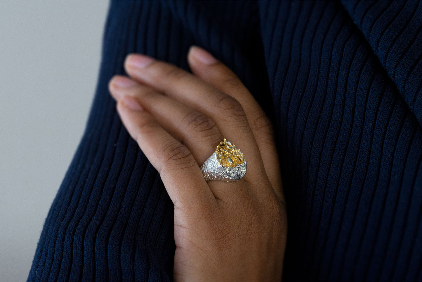A close-up of a hand wearing a large statement sea silver ring with a yellow center with a white sun and a textured silver band, set against a dark navy ribbed sweater.