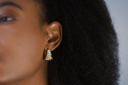 A close-up side profile of a person with dark curly hair wearing a small pair of gold vermeil and silver earrings with a pair of sculptural gold vermeil and silver earrings featuring two dolphins leaping out of the sea, mid-splash. 