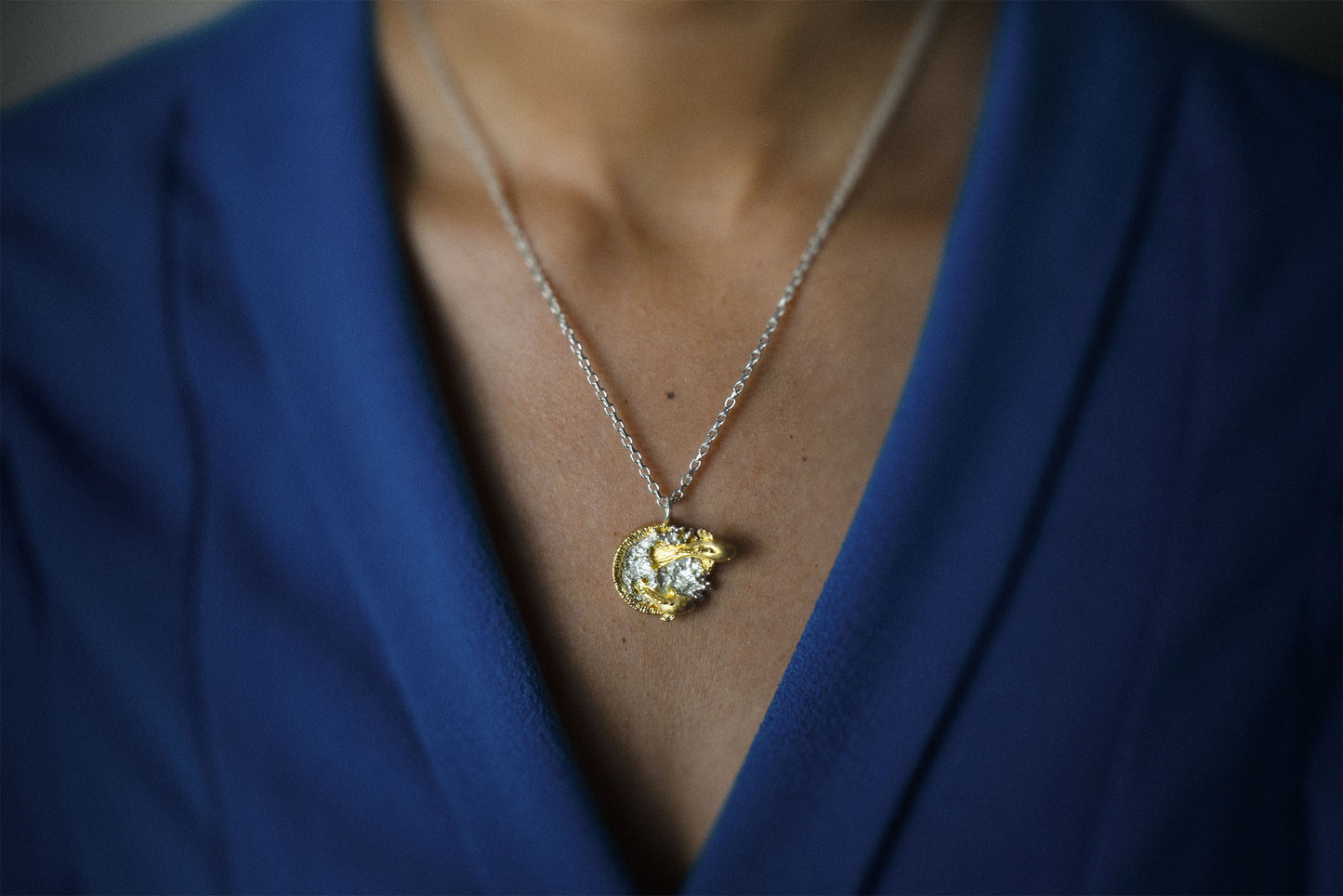 A close-up shot of a person wearing a delicate gold pendant necklace with a clear crystal center, styled against a royal blue blazer.