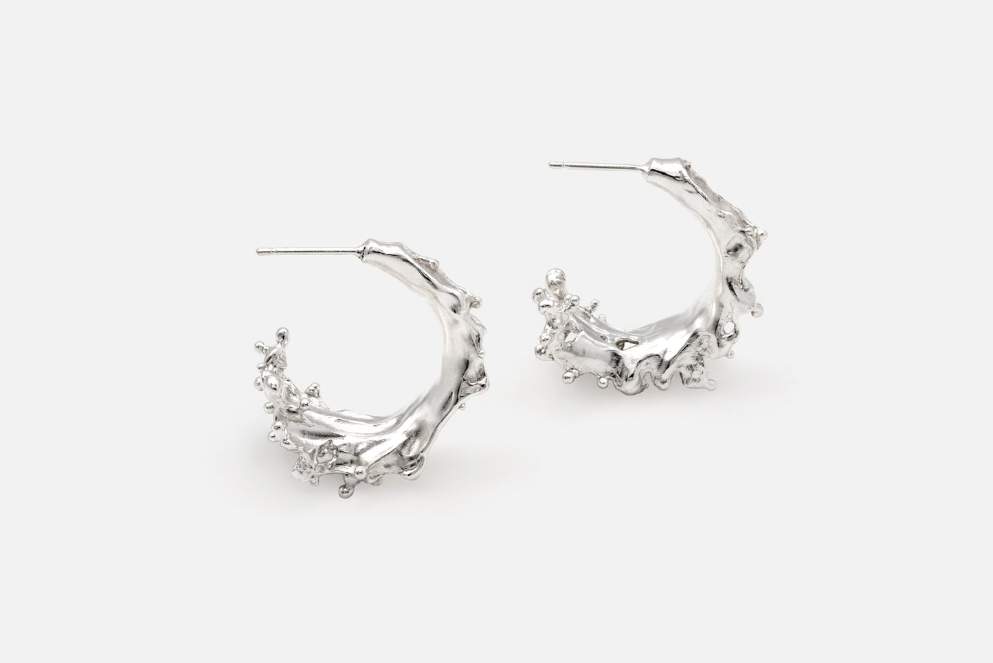 A pair of silver crescent-shaped hoop earrings with textured, organic details like the shape of sea waves rising at their highest point, shown on a light gray background.