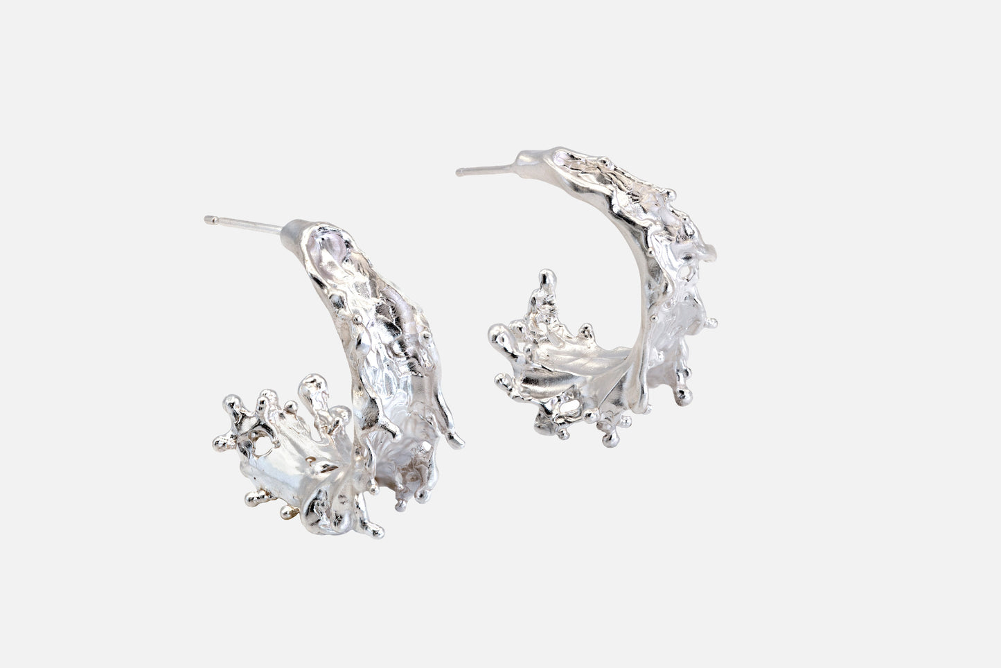 A pair of silver crescent-shaped hoop earrings with textured, organic details like the shape of sea waves rising at their highest point, shown on a light gray background.
