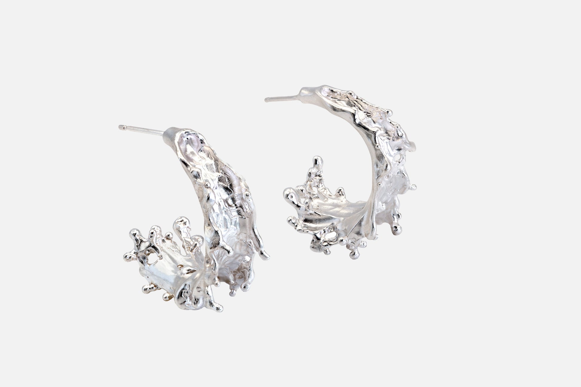 A pair of silver crescent-shaped hoop earrings with textured, organic details like the shape of sea waves rising at their highest point, shown on a light gray background.