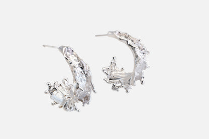A pair of silver crescent-shaped hoop earrings with textured, organic details like the shape of sea waves rising at their highest point, shown on a light gray background.
