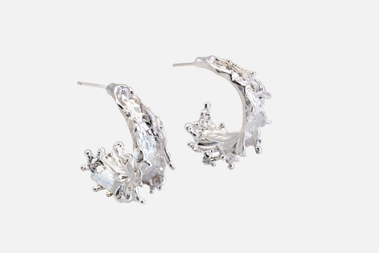 A pair of silver crescent-shaped hoop earrings with textured, organic details like the shape of sea waves rising at their highest point, shown on a light gray background.