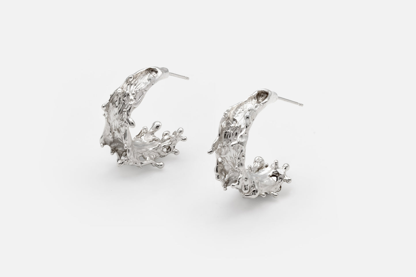 A pair of silver crescent-shaped hoop earrings with textured, organic details like the shape of sea waves rising at their highest point, shown on a light gray background.