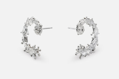 A pair of silver earrings with a textured, organic design like the waters of the sea swirling in a circular pattern, displayed on a clean light gray background.