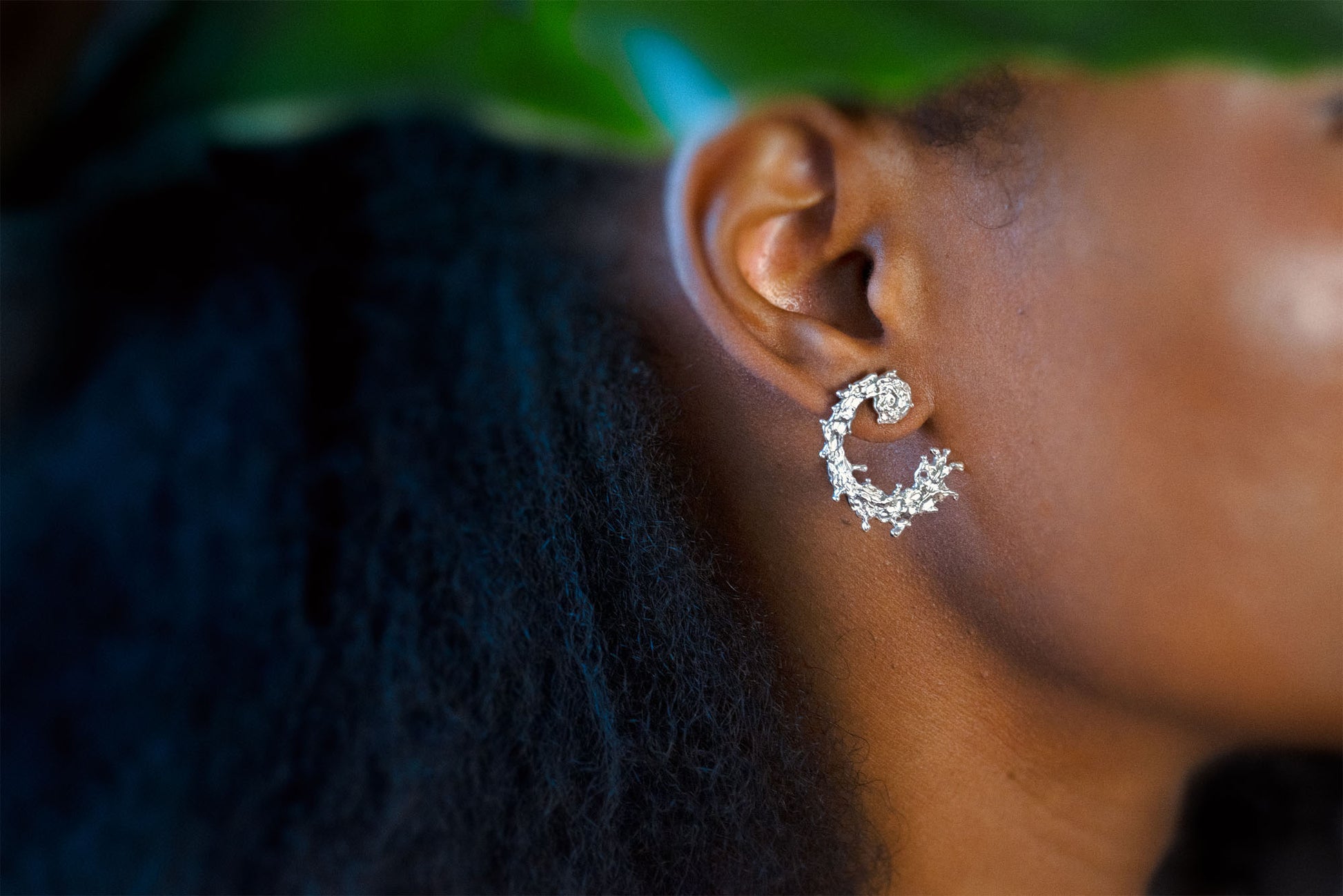 A close-up side profile of a person wearing a silver earring with a textured, organic design like the waters of the sea swirling in a circular pattern, displayed on a clean light gray background.