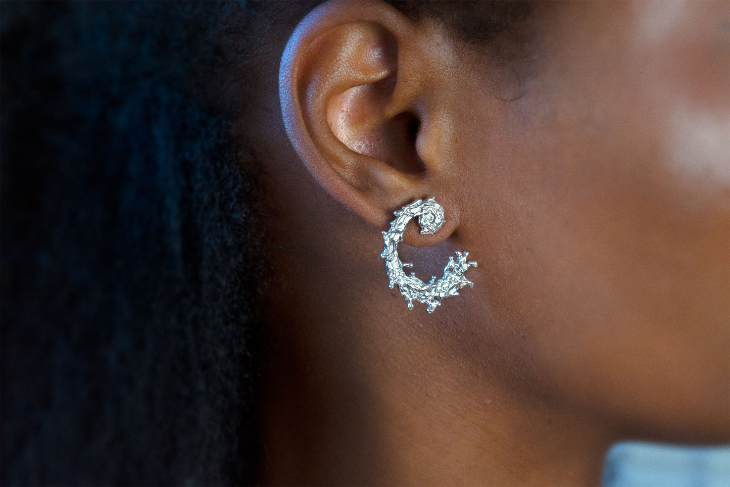 A close-up side profile of a person wearing a silver earring with a textured, organic design like the waters of the sea swirling in a circular pattern, displayed on a clean light gray background.