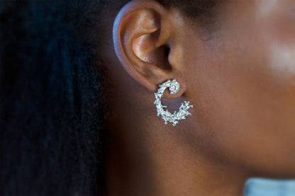 A close-up side profile of a person wearing a silver earring with a textured, organic design like the waters of the sea swirling in a circular pattern, displayed on a clean light gray background.