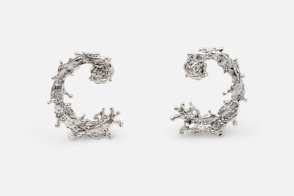 A pair of silver earrings with a textured, organic design like the waters of the sea swirling in a circular pattern, displayed on a clean light gray background.
