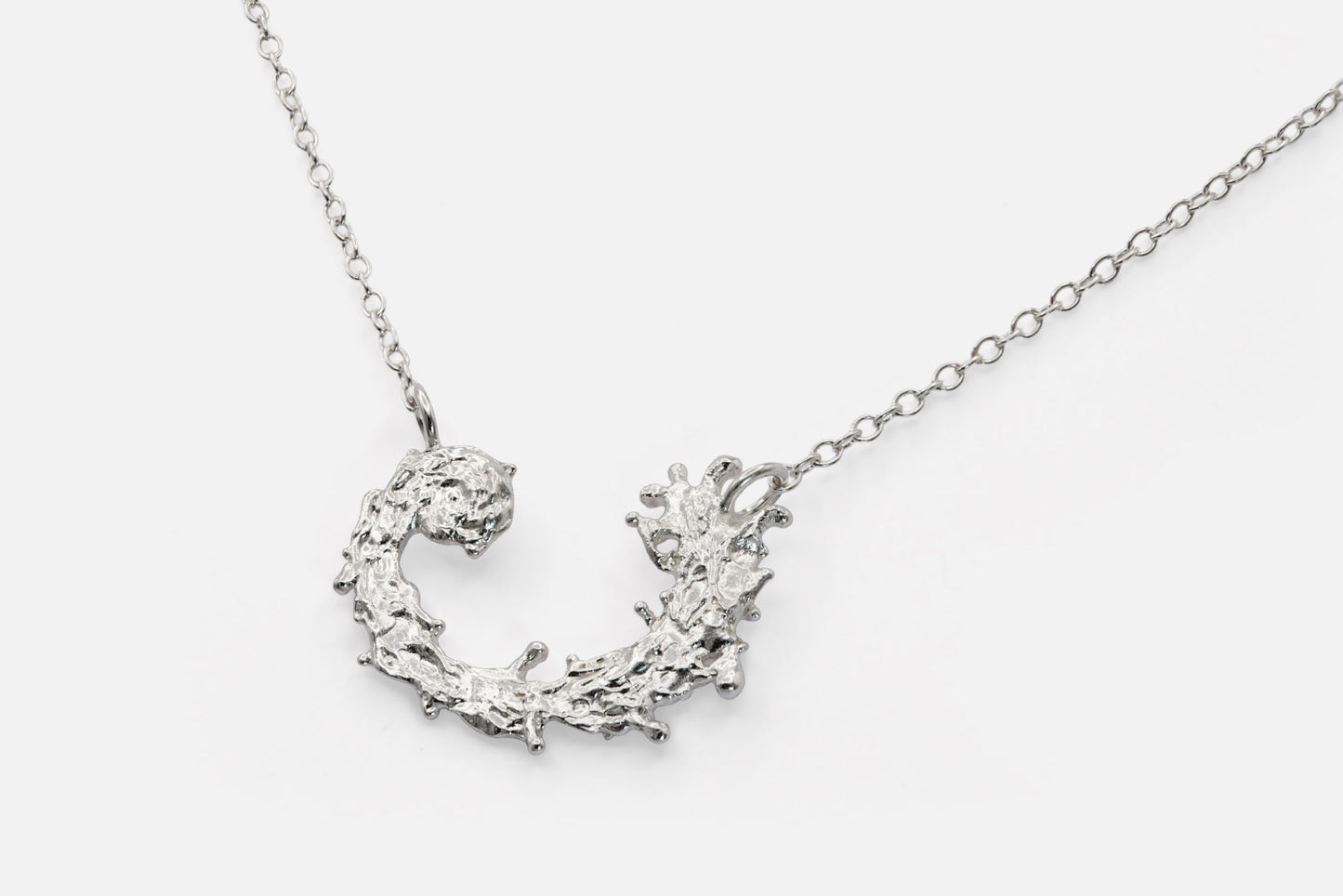 A silver necklace featuring a textured, organic crescent moon pendant with a design like the waters of the sea swirling in a circular pattern, suspended from a delicate link chain against a light gray background.
