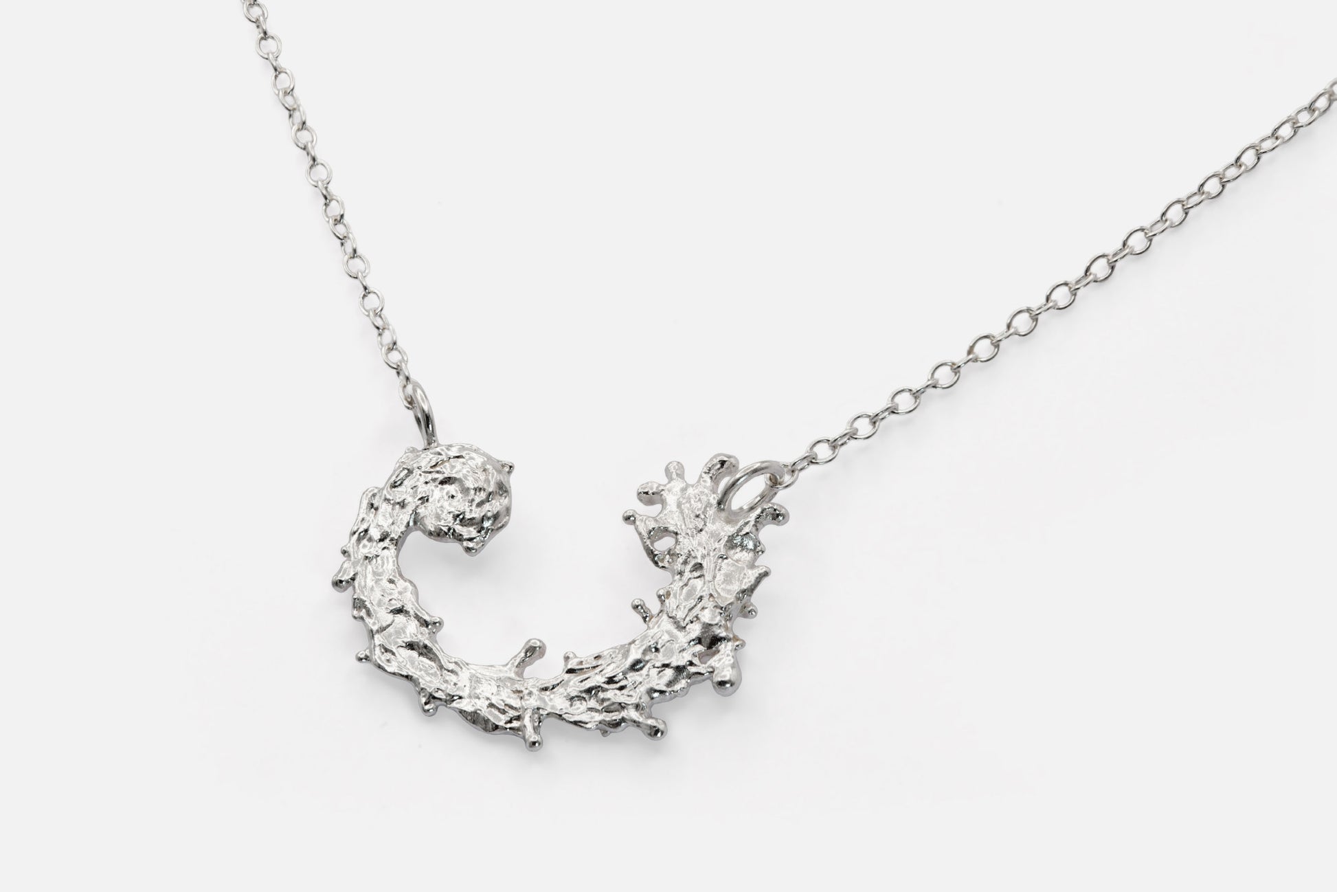 A silver necklace featuring a textured, organic crescent moon pendant with a design like the waters of the sea swirling in a circular pattern, suspended from a delicate link chain against a light gray background.