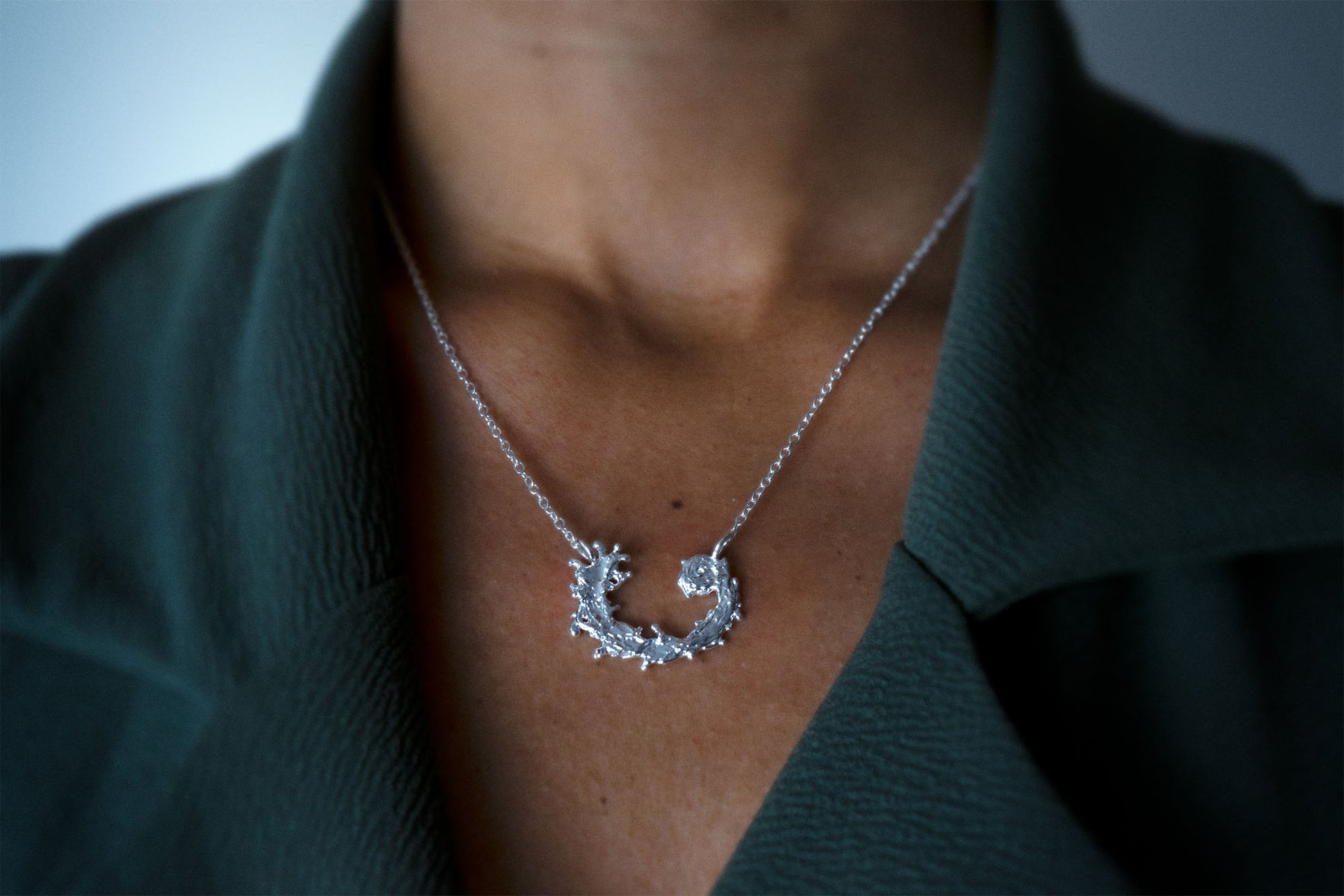 Woman wearing a silver necklace featuring a textured, organic crescent moon pendant with a design like the waters of the sea swirling in a circular pattern, set against a dark green jacket.