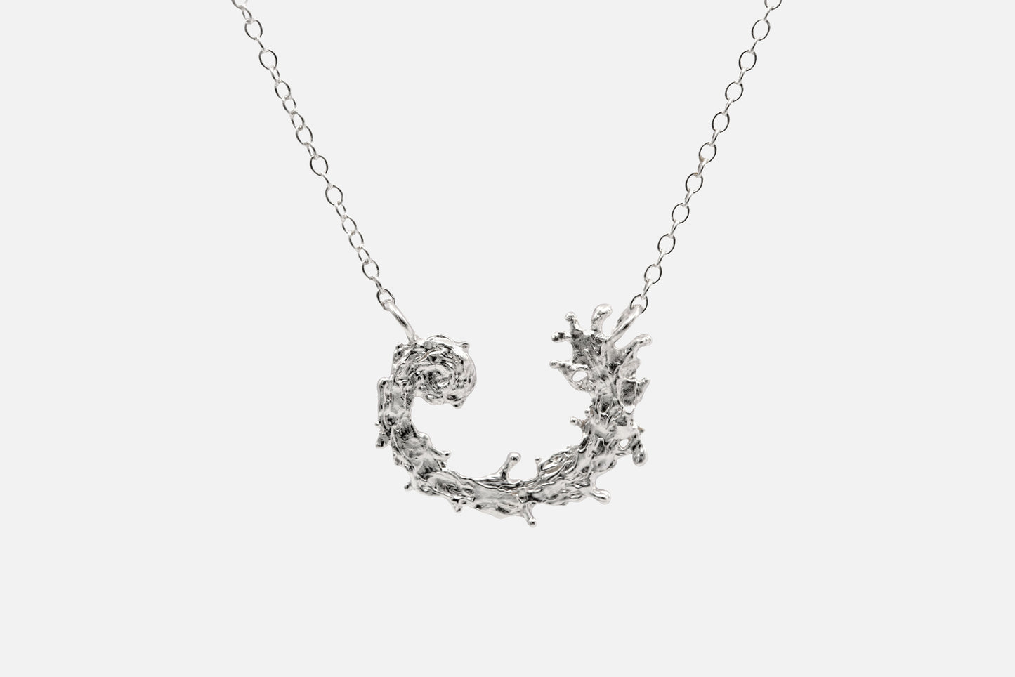 A silver necklace featuring a textured, organic crescent moon pendant with a design like the waters of the sea swirling in a circular pattern, suspended from a delicate link chain against a light gray background.