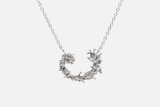 A silver necklace featuring a textured, organic crescent moon pendant with a design like the waters of the sea swirling in a circular pattern, suspended from a delicate link chain against a light gray background.