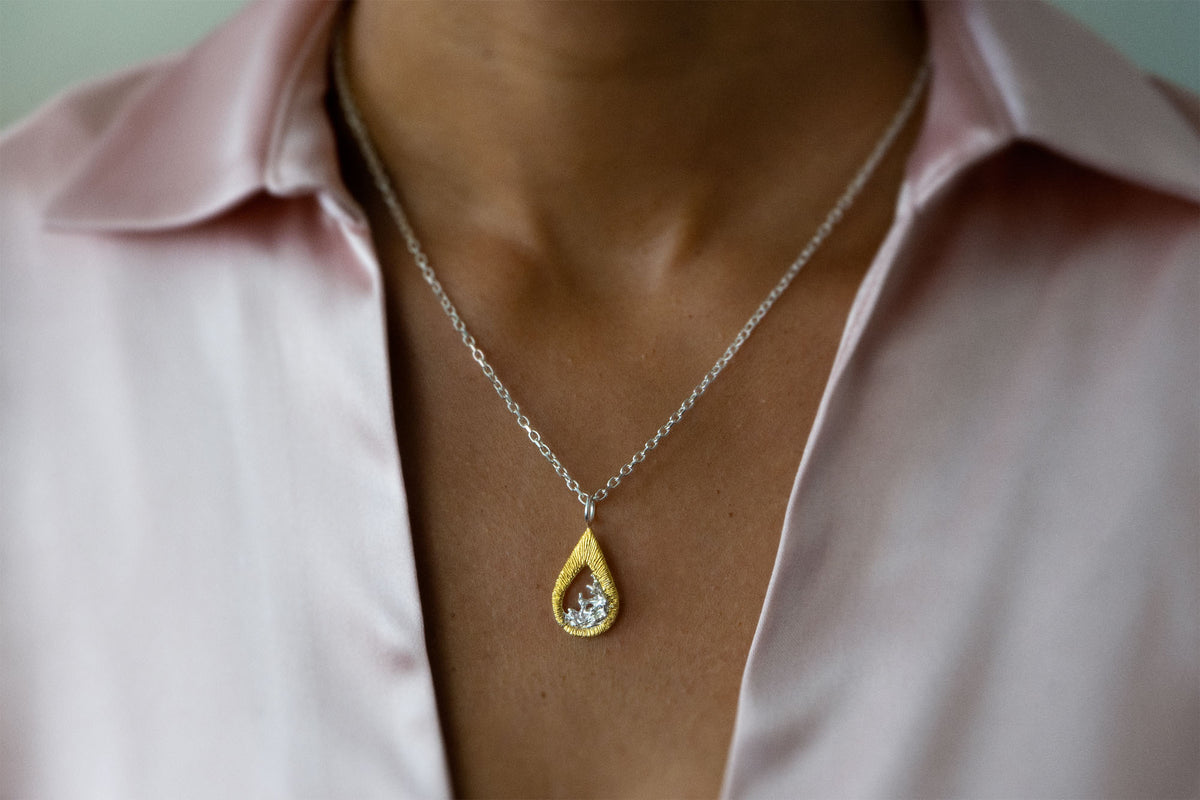 A close-up of a person wearing a delicate silver chain necklace with a teardrop-shaped gold vermeil and silver pendant necklace featuring a textured gold outer frame and a silver-toned inner design shaped like moving water, suspended from a silver chain, styled with a light pink satin shirt.
