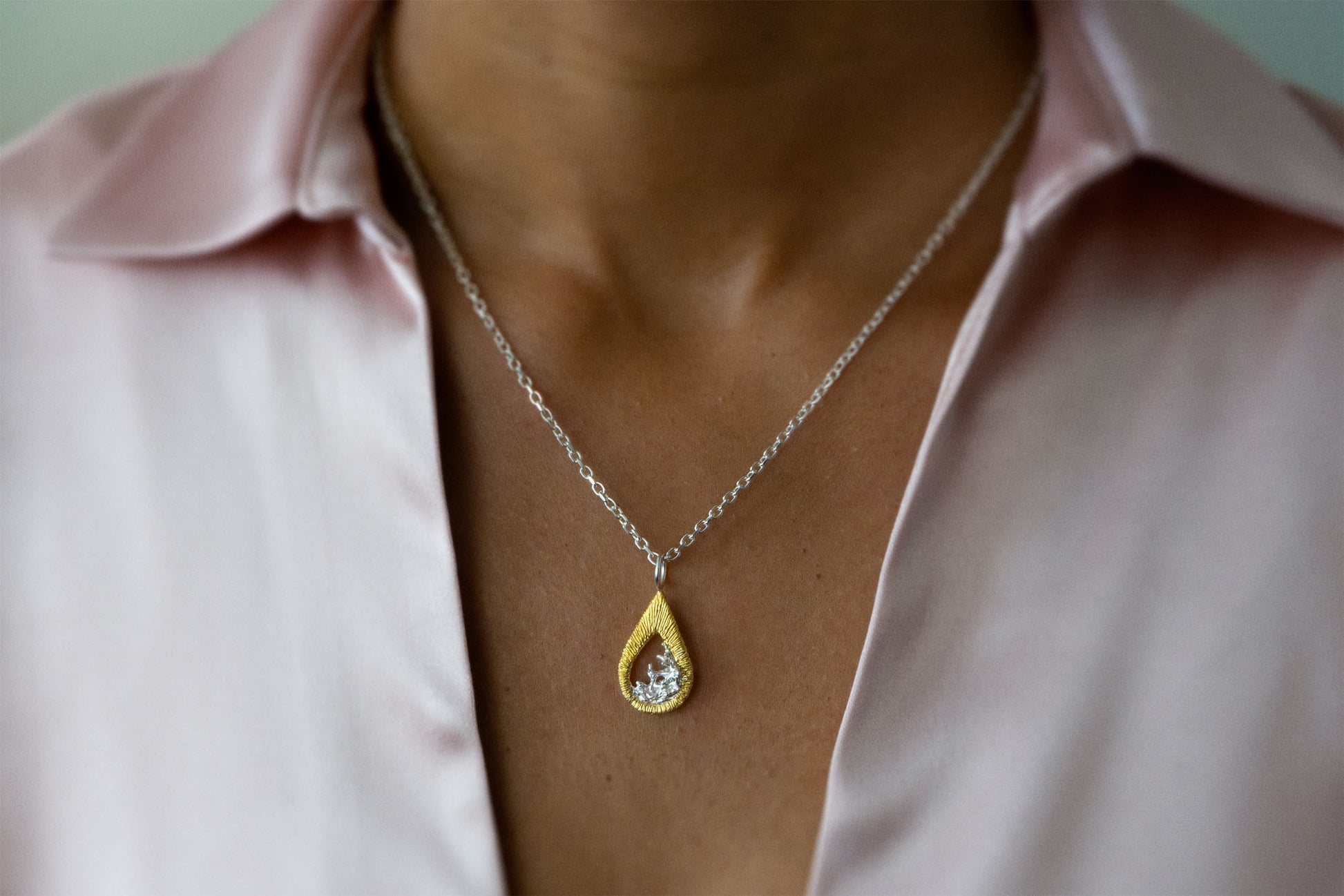 A close-up of a person wearing a delicate silver chain necklace with a teardrop-shaped gold vermeil and silver pendant necklace featuring a textured gold outer frame and a silver-toned inner design shaped like moving water, suspended from a silver chain, styled with a light pink satin shirt.