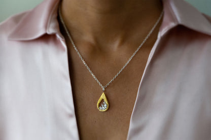 A close-up of a person wearing a delicate silver chain necklace with a teardrop-shaped gold vermeil and silver pendant necklace featuring a textured gold outer frame and a silver-toned inner design shaped like moving water, suspended from a silver chain, styled with a light pink satin shirt.