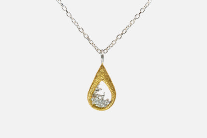 A teardrop-shaped gold vermeil and silver pendant necklace featuring a textured gold outer frame and a silver-toned inner design shaped like moving water, suspended from a silver chain against a light gray background.