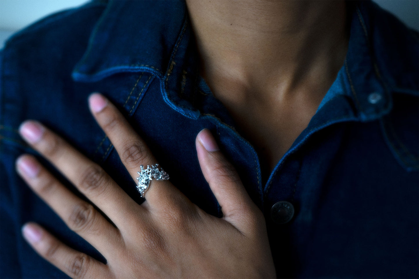A close-up shot of a person wearing a silver ring with a sculptural, textured top, like waves of the sea rising to meet, with their hand resting on their denim shirt.