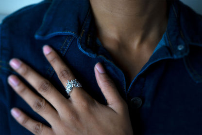 A close-up shot of a person wearing a silver ring with a sculptural, textured top, like waves of the sea rising to meet, with their hand resting on their denim shirt.