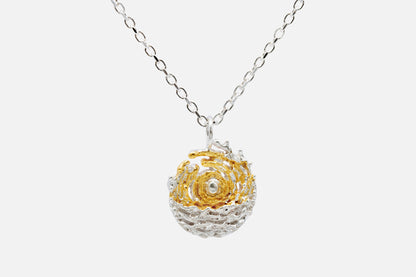 A delicate silver chain necklace featuring a spherical pendant with a gold swirl around a central sun, set against a textured silver background like the sea and tides moving at dawn, shown against a light gray background.